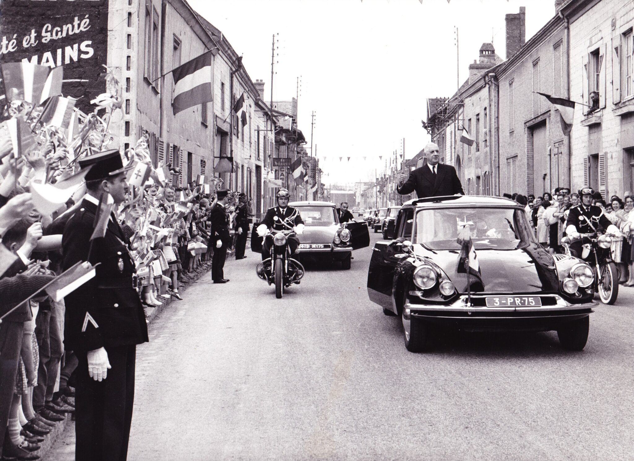 Trente Glorieuses : cortège officiel dans une rue française, berline DS, foule et drapeaux.