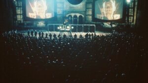 Scène inspirée de 1984 montrant une foule devant un écran géant diffusant le visage de Big Brother dans un décor totalitaire.