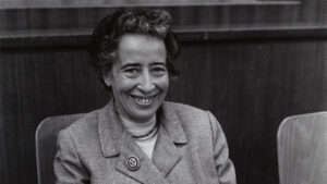 Portrait en noir et blanc d’Hannah Arendt souriant, assise en tailleur-pantalon et veste, figure majeure de la pensée politique.